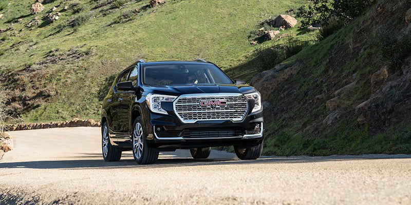 A 2024 GMC Terrain driving on a road surrounded by trees and clear blue sky.