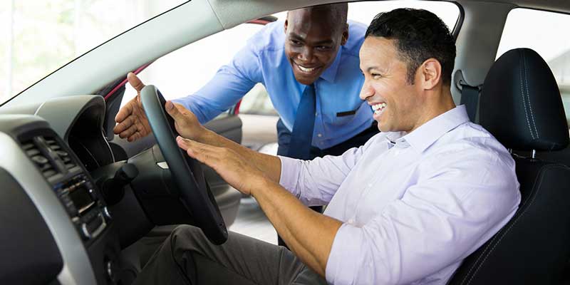 Two men in a car, smiling and pointing at the steering wheel, with one man appearing to assist the other in a sale.